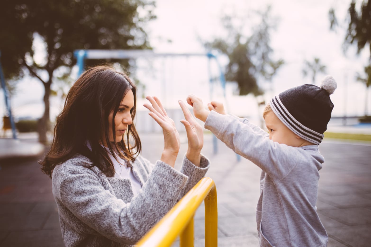 A nanny playing a clapping game with a toddler at a playground